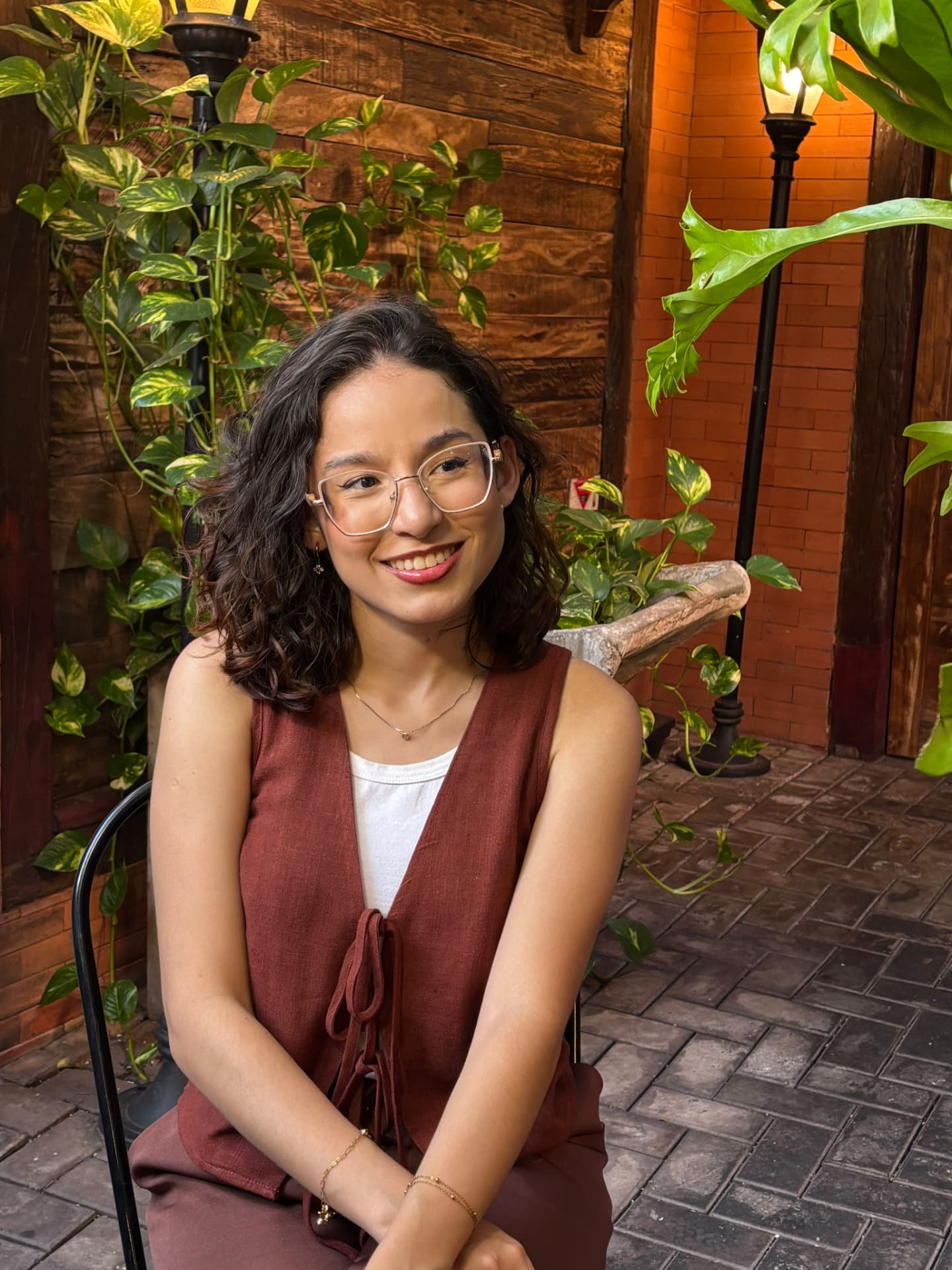 Thafnes Samara sorrindo, segurando um livro em um ambiente com plantas e luz quente